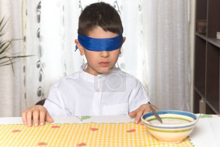Einem achtjährigen kaukasischen Jungen, der zu Hause am Tisch sitzt, werden mit ernster Geste die Augen verbunden. Das Kind hat gerade einen blinden Geschmackstest eines Lebensmittels gemacht.