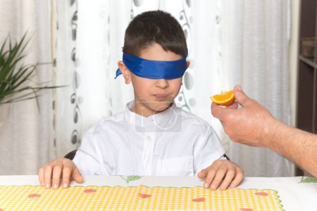 Blindfolded 8-year-old boy testing the taste of a piece of orange being given by a person, apart from the boy, you can also see the arm of an adult who is holding the fruit.
