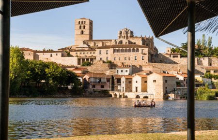 View of the cathedral with the mills and the Duero river where there is a barge crossing the water with tourists during a sunny summer day in Zamora, Spain. 