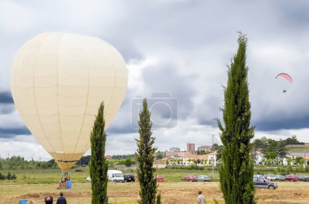 Photo for Hot air balloon on the ground of a field and paramotor flying in the sky at a leisure event next to the city. - Royalty Free Image