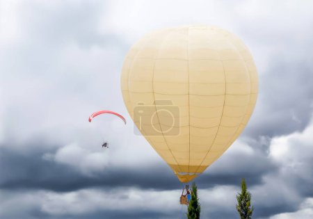 Photo for Large hot air balloon flying in the sky with clouds next to a paramotor at an air festival. - Royalty Free Image