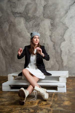 Foto de Hermosa joven posando en estudio con vestido blanco, chaqueta negra y sombrero gris - Imagen libre de derechos