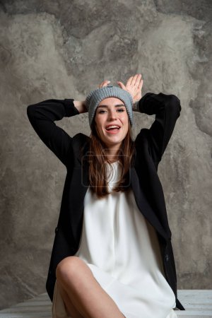 Téléchargez les photos : Belle jeune femme posant en studio en robe blanche, veste noire et chapeau gris - en image libre de droit