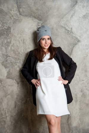 Téléchargez les photos : Belle jeune femme posant en studio en robe blanche, veste noire et chapeau gris - en image libre de droit