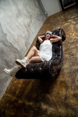 Téléchargez les photos : Jeune belle femme posant sur canapé en robe blanche et chapeau gris - en image libre de droit