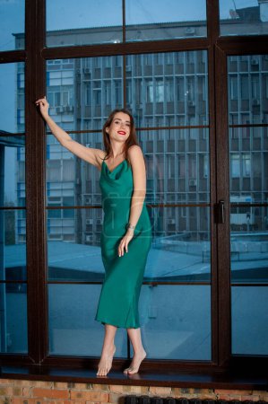 Foto de Joven hermosa mujer vistiendo vestido verde, posando cerca de ventana panorámica - Imagen libre de derechos