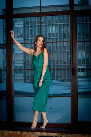 Photo for Young beautiful woman wearing green dress, posing near panoramic window - Royalty Free Image