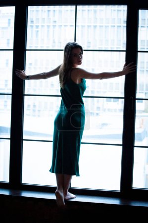 Téléchargez les photos : Jeune belle femme portant une robe verte, posant près de la fenêtre panoramique - en image libre de droit