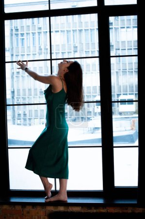 Téléchargez les photos : Jeune belle femme portant une robe verte, posant près de la fenêtre panoramique - en image libre de droit