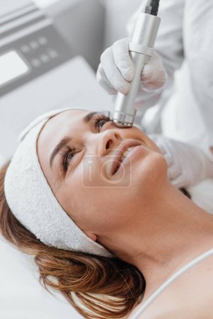 Téléchargez les photos : Electroporation without injection mesotherapy. Woman cosmetologist performs cosmetic procedure on skin of the face to reduce wrinkles, restore, regenerate and rejuvenate the girls face. Beauty clinic - en image libre de droit