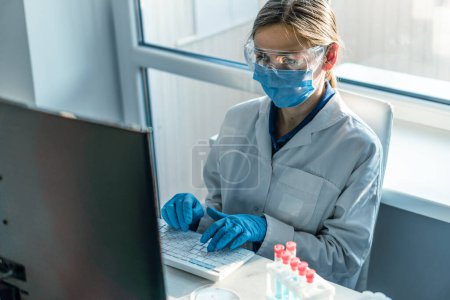 Téléchargez les photos : Une chercheuse est assise dans un laboratoire, derrière un écran d'ordinateur personnel. Dans le contexte des statistiques de recherche. Travailleur médical pharmaceutique en protection travaille au clavier. - en image libre de droit