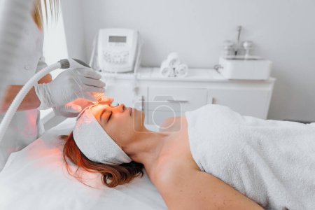 Radiofrequency face lifting. The beautician performs the procedure in the beauty salon using an LED device. Hardware anti-aging procedure. Young woman lying resting and relaxing. High quality photo