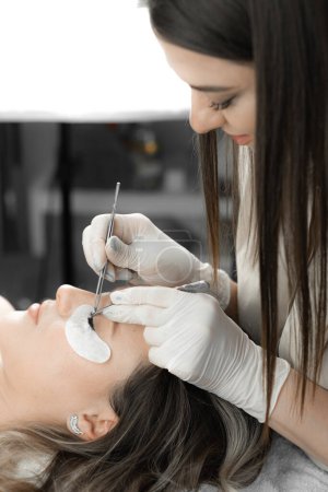 The woman lies relaxed, and the lashmaker carefully builds eyelashes, creating a wonderful decoration effect for her face and emphasizing her beauty. Cosmetic procedure in the beauty salon is done by