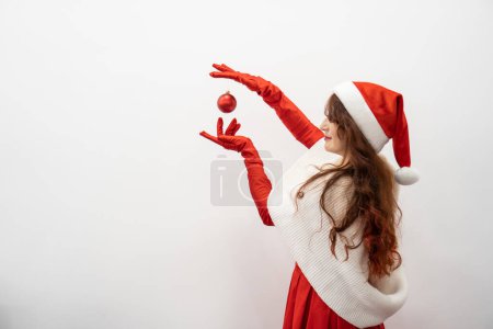 Téléchargez les photos : Un ballon rouge dans les mains dans les rumeurs rouges d'une femme heureuse en bonnet Santas apporte une humeur instantanée de joie et d'émerveillement à la maison. Femme en rouge sur fond blanc isolé. Photo de haute qualité - en image libre de droit