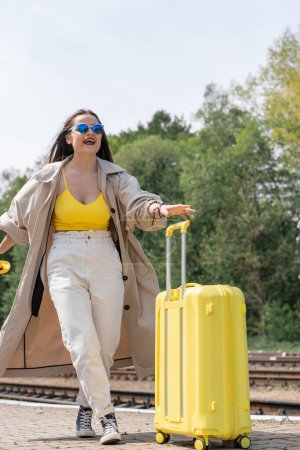 Téléchargez les photos : Parcours d'aventure : une femme en jaune avec une valise est prête à découvrir où son nouveau chemin mènera. Lumière du futur : l'image d'un voyageur en lunettes de soleil et vêtements jaunes incarne la - en image libre de droit