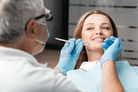 Eine Patientin im Zahnarztstuhl zeigt freudig ihr schönes Lächeln, das dank der Behandlung durch einen Zahnarzt entstanden ist. Der Arzt, der den Eingriff durchgeführt hat, ist mit dem Aussehen zufrieden und bemüht sich,