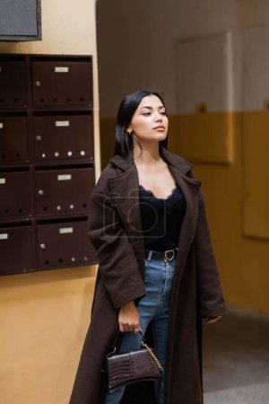 Photo for Brunette woman in trendy coat standing with handbag near building with mailboxes in prague - Royalty Free Image