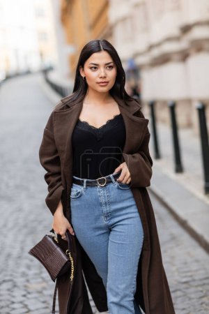 Téléchargez les photos : Femme brune à la mode avec sac à main touchant ceinture de jeans sur rue floue dans la prague - en image libre de droit
