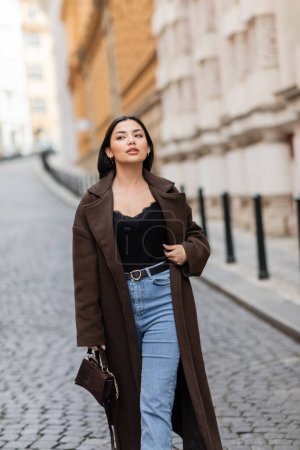 trendige Frau in braunem Mantel, die mit Handtasche auf der Prager Straße spaziert und wegschaut