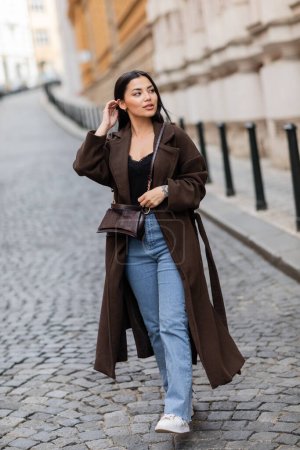 brunette woman in trendy coat with crossbody fixing hair and looking away on blurred street in prague