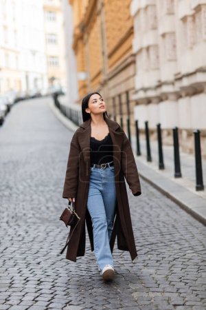 volle Länge der Frau in stylischem Mantel und Jeans, die in Prag spazieren geht und wegschaut