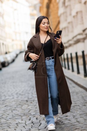 Téléchargez les photos : Brunette femme en manteau et jeans regardant smartphone tout en marchant dans la rue dans la prague - en image libre de droit