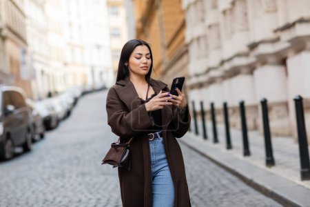 trendige brünette Frau mit Crossbody mit Handy auf verschwommener Straße in Prag