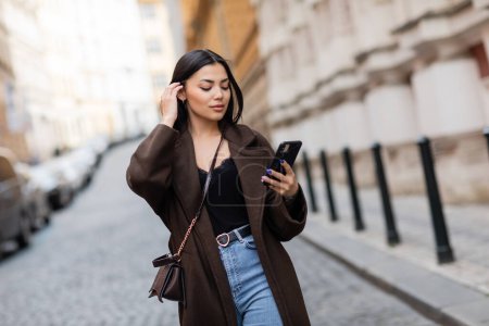 Photo for Young woman in brown coat fixing hair and looking at cellphone on blurred street in prague - Royalty Free Image