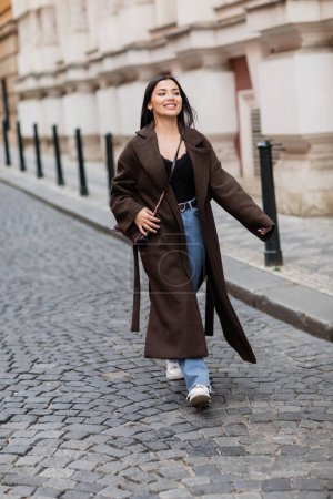 full length of excited woman in stylish coat walking on pavement in prague