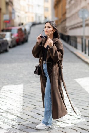 volle Länge der hübschen brünetten Frau im Mantel mit Sonnenbrille auf der Straße in Prag