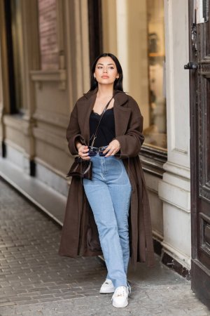 full length of young woman in stylish coat and jeans holding sunglasses near building in prague