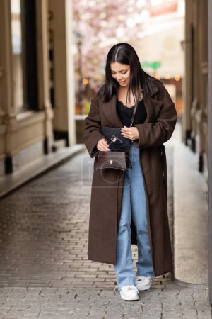 trendige brünette Frau nimmt Handy aus Kreuzkörper auf der Straße in Prag