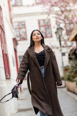 hübsche brünette Frau in trendigem Mantel und drahtlosen Kopfhörern, die mit Handtasche in Prag spazieren geht