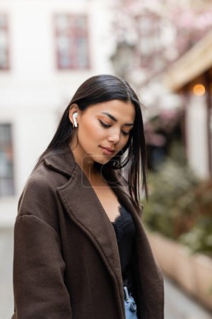 Téléchargez les photos : Jeune femme brune dans un manteau élégant écoutant de la musique dans un écouteur sans fil dans la rue à Prague - en image libre de droit