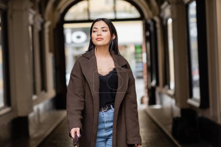 Téléchargez les photos : Femme élégante en manteau marron et jeans regardant loin près de la voûte de bâtiment à prague - en image libre de droit