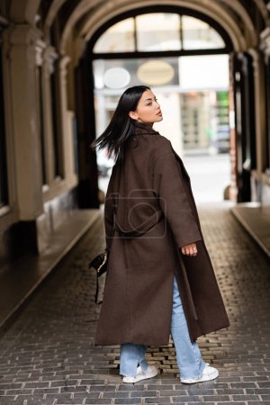 back view of young brunette woman in brown coat looking away under arch building in prague