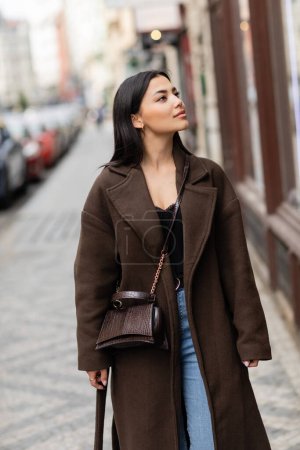 Téléchargez les photos : Jolie femme brune en manteau marron avec bandoulière tendance regardant loin sur la rue urbaine dans la prague - en image libre de droit