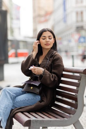 stylische brünette Frau mit Kopfhörertasche sitzt mit geschlossenen Augen auf Bank in Prag 