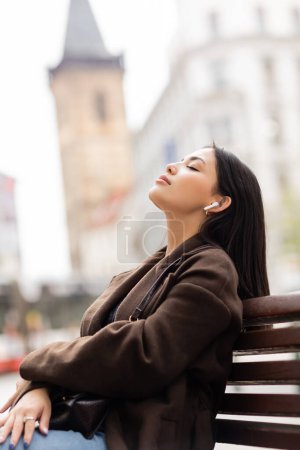 Téléchargez les photos : Vue latérale de la femme en manteau d'automne relaxant sur le banc tout en écoutant de la musique dans un écouteur sans fil dans la rue à Prague - en image libre de droit