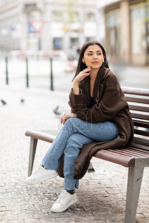 hübsche Frau in trendiger Herbstkleidung hört Musik in drahtlosen Kopfhörern auf Bank in Prag und schaut weg