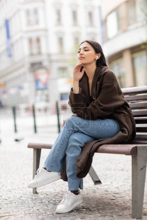 dreamy woman sitting on bench in prague and listening music in wireless earphone