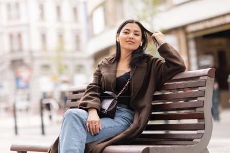 Photo for Happy brunette woman touching hair and listening music in wireless earphones on bench in prague - Royalty Free Image