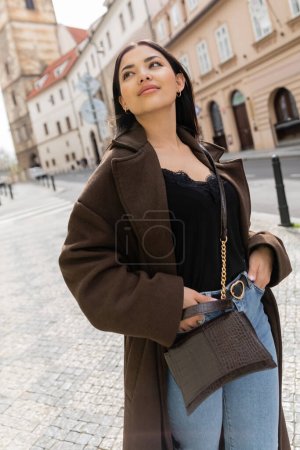 young woman in trendy coat standing with hands in pockets of jeans and looking away in prague 