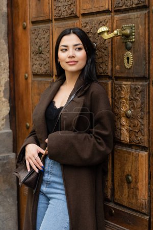 Lächelnde Frau im trendigen Mantel mit Kreuzkörper lehnt an geschnitzter Tür und schaut in Prag weg