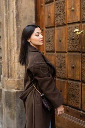 brünette Frau in trendigem braunem Mantel mit Crossbody steht neben geschnitzter Holztür in Prag