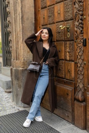smiling woman in jeans and coat touching hair near carved wooden door in prague