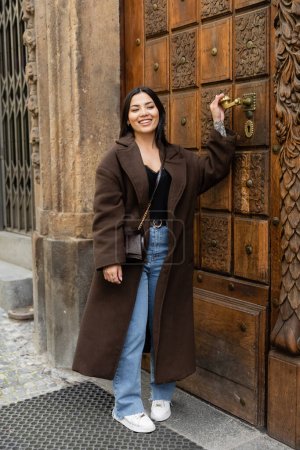 happy brunette woman in trendy coat touching door handle near building in prague