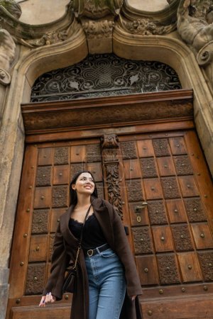 low angle view of brunette woman in jeans and trendy coat smiling near carved wooden door in prague 