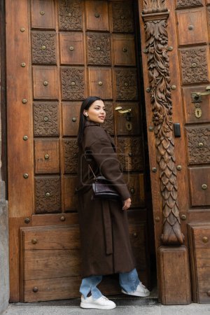 Frau in Mantel und drahtlosem Kopfhörer schaut in der Nähe der geschnitzten Tür in Prag weg