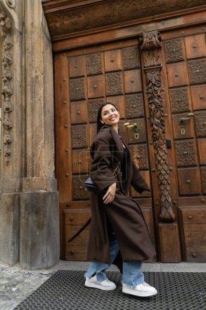 fröhliche Frau im trendigen braunen Mantel, die in Prag nahe der geschnitzten Holztür spaziert und wegschaut